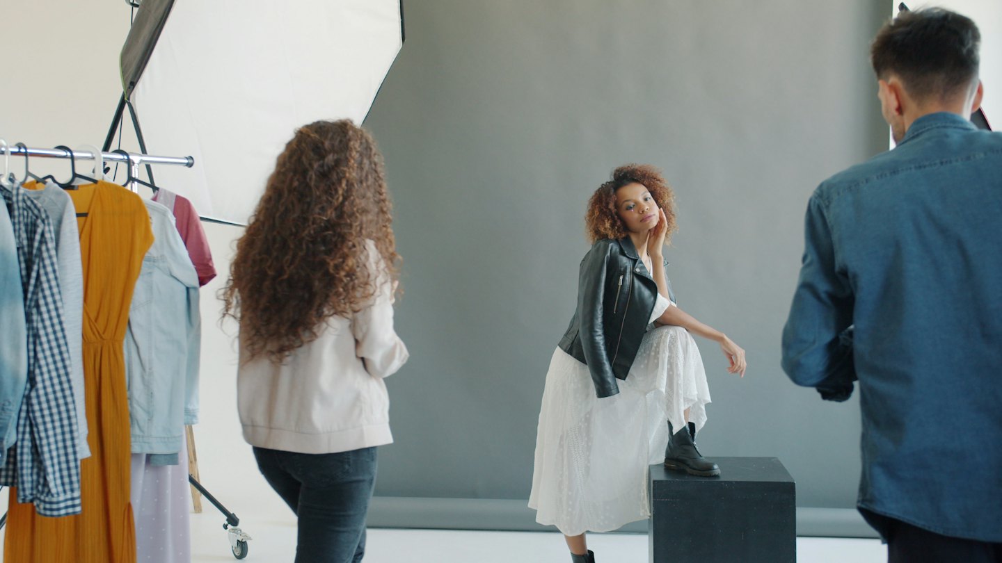Model poses in studio with photographer and stylist nearby.